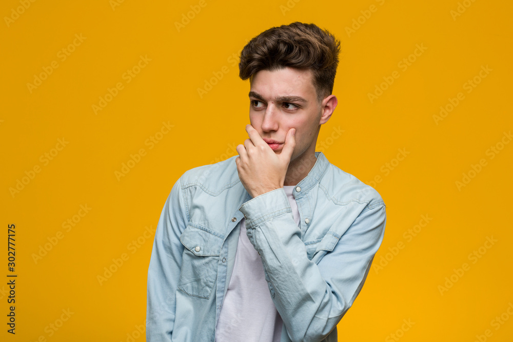 Young handsome student wearing a denim shirt thoughtful looking to a copy space covering mouth with hand.