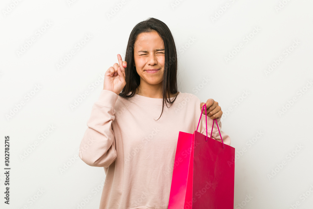 Young hispanic woman holding a shopping bag crossing fingers for having luck Young hispanic woman holding a shopping bag crossing fingers for having luck