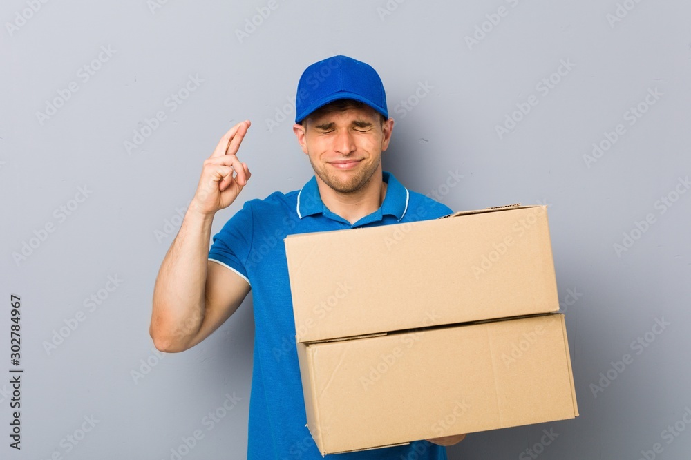 Young man delivering packages crossing fingers for having luck Young man delivering packages crossing fingers for having luck