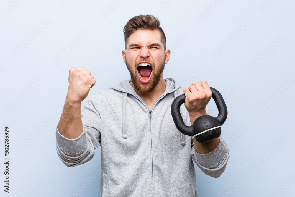 Young caucasian sportsman holding a dumbbell cheering carefree and excited. Victory concept. - obrazy, fototapety, plakaty 