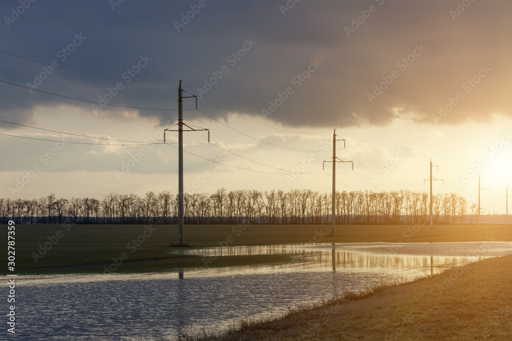 Power line along the road and green field with large puddles after rain in early spring