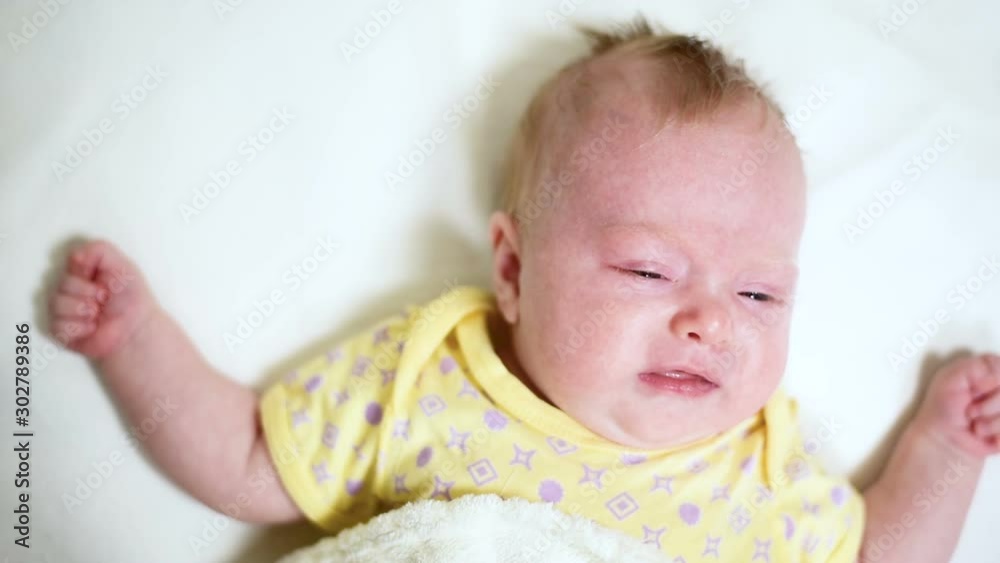 Crying blond baby lies on a white bed. Close up Crying blond baby lies on a white bed. Close up