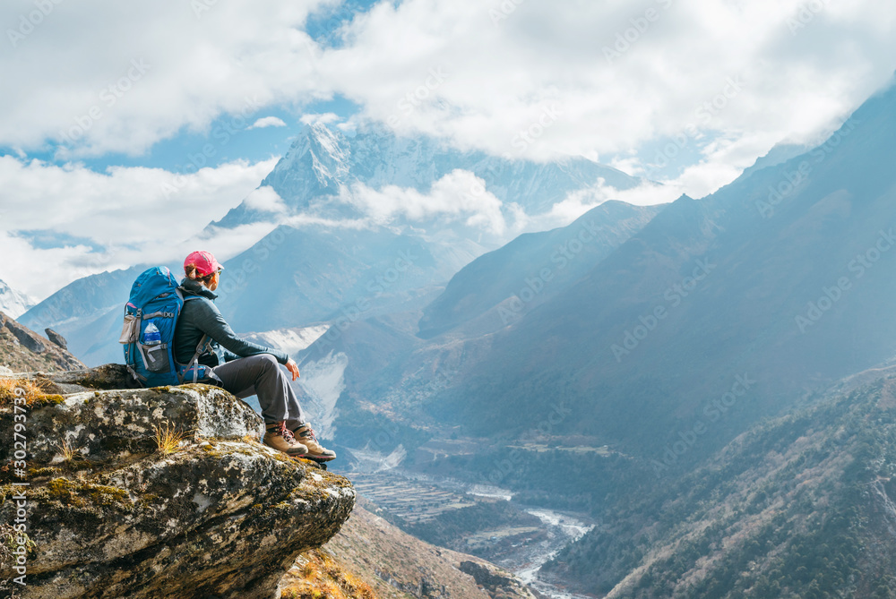 Young hiker backpacker female sitting on the cliff edge and enjoying Ama Dablam 6,812m peak view during Everest Base Camp (EBC) trekking route near Phortse, Nepal. Active vacations concept image