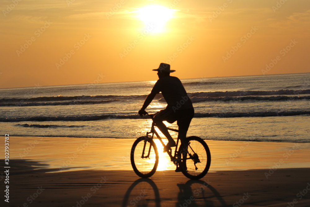 Fototapeta premium Man silhouette bycicling on the beach during the sunset. Golden sky with the sun in the sky. Riviera beach, Bertioga, Brazil