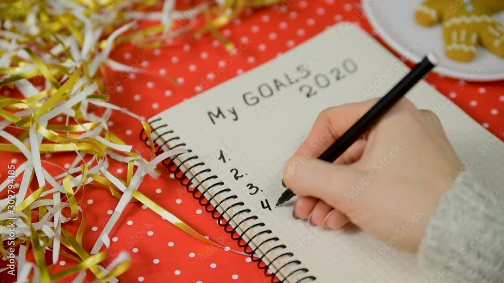 Female hands writing goals on 2020 in a notebook. Red background and tinsel. New Years concept