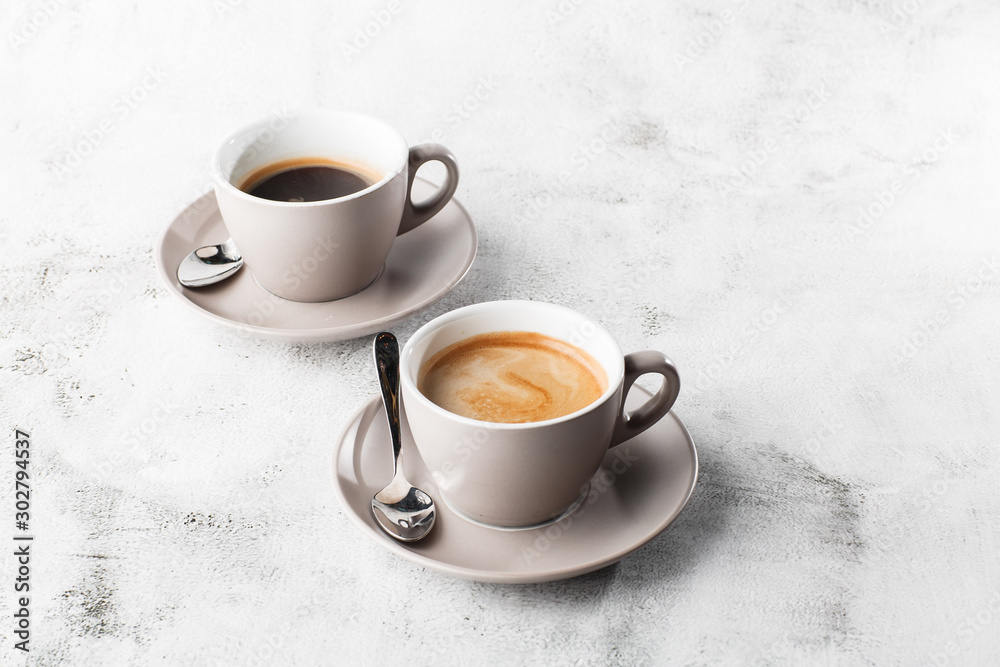 Two white cups of hot black coffee with milk isolated on bright marble background. Overhead view, copy space. Advertising for cafe menu. Coffee shop menu. Horizontal photo. Two white cups of hot black coffee with milk isolated on bright marble background. Overhead view, copy space. Advertising for cafe menu. Coffee shop menu. Horizontal photo.