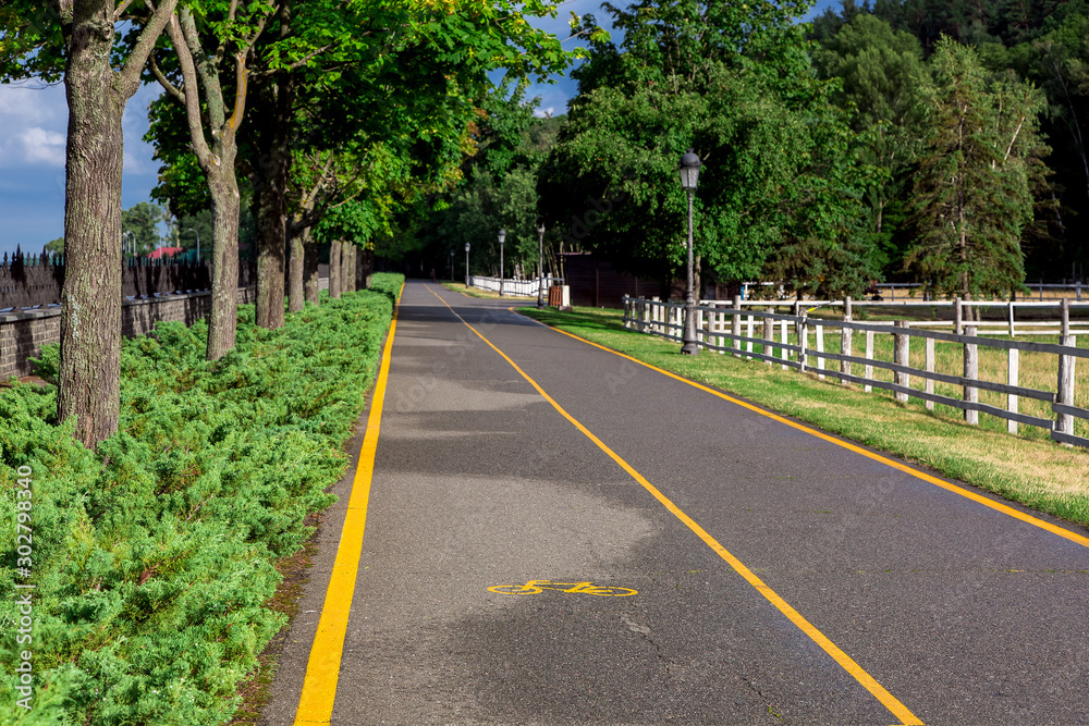 asphalt road with yellow markings and a bicycle symbol for the lane of bikes in the park on a sunny summer day on the side of the bushes with green trees and a wooden pasture fence.