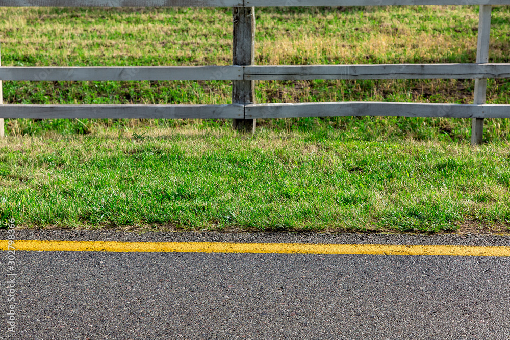 roadside with yellow marking with green grass and in the background a wooden pasture fence on a sunny summer day.