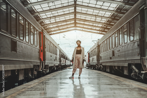 African woman in a white cloak and spectacles is standing in the center of a station between two highway trains waiting for departure at the platform indoors of a railroad depot; a triangle roof atop