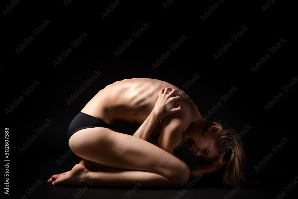 Fototapeta premium Helpless woman position embryo posing on a black isolated background. The concept of helplessness and loneliness. Female fear and hopelessness before fear and unchartedness