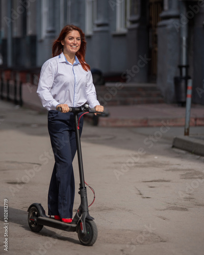 A red-haired girl in a white shirt drives an electric scooter along the wall. A business woman in a trouser suit and red high heels rides around the city in a modern car. dress code in the office.