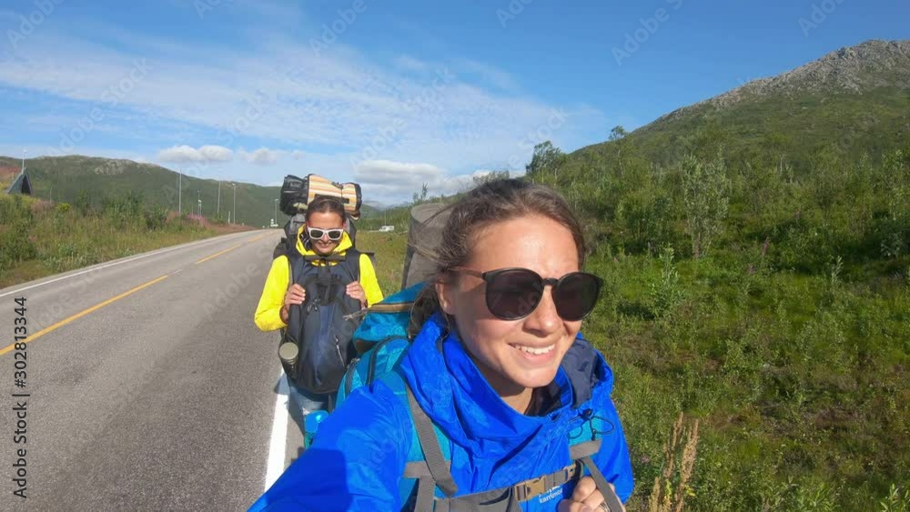 Two young women travel bloggers walking a Norwegian road with heavy backpacks and taking a selfie. Two young women travel bloggers walking a Norwegian road with heavy backpacks and taking a selfie.