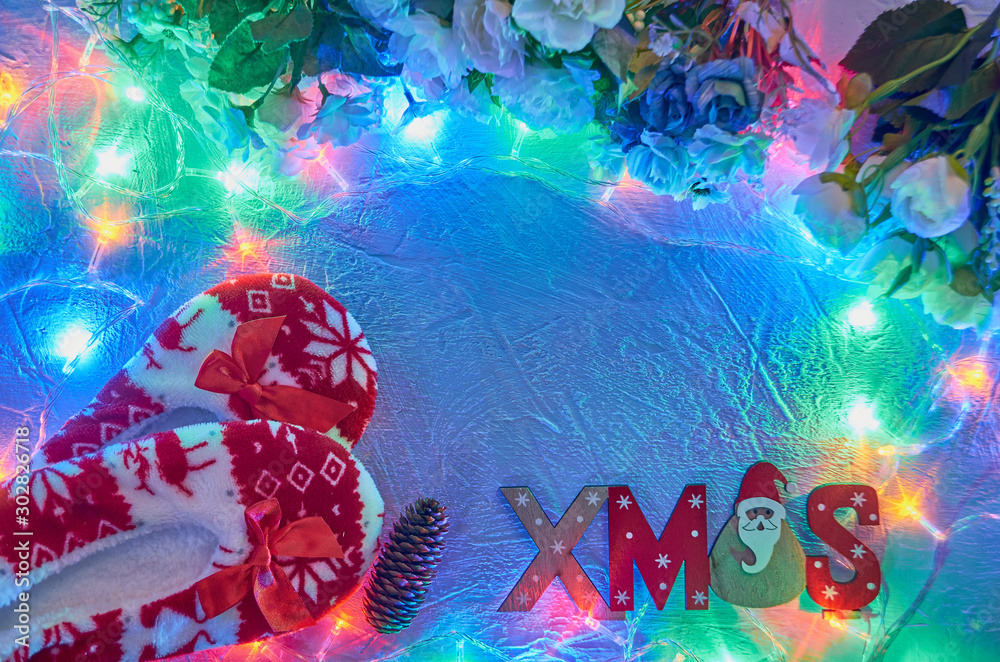 Obraz Świąteczne tło uroczysty z miejscem na tekst. Kwiaty, girlandy, drewniany napis z Mikołajem i ciepłe czerwone świąteczne kapcie z wzorem jelenia.