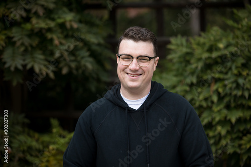 A caucasian male wearing a black hoodie sweater and glasses poses in a garden while surrounded by fall foliage on a cold afternoon.