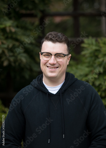 A caucasian male wearing a black hoodie sweater and glasses poses in a garden while surrounded by fall foliage on a cold afternoon.