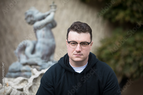 A caucasian male wearing a black hoodie sweater and glasses poses in a garden while surrounded by fall foliage on a cold afternoon.