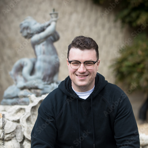 A caucasian male wearing a black hoodie sweater and glasses poses in a garden while surrounded by fall foliage on a cold afternoon.