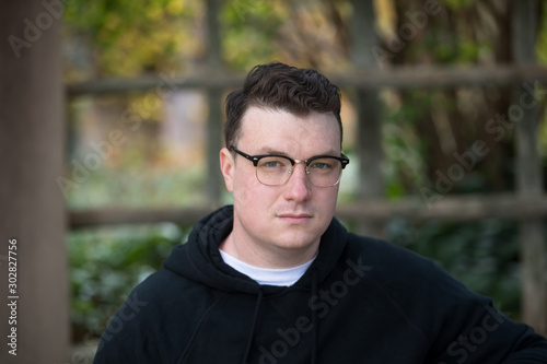 A caucasian male wearing a black hoodie sweater and glasses poses in a garden while surrounded by fall foliage on a cold afternoon.