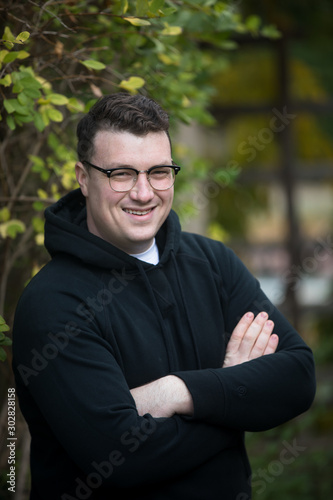 A caucasian male wearing a black hoodie sweater and glasses poses in a garden while surrounded by fall foliage on a cold afternoon.