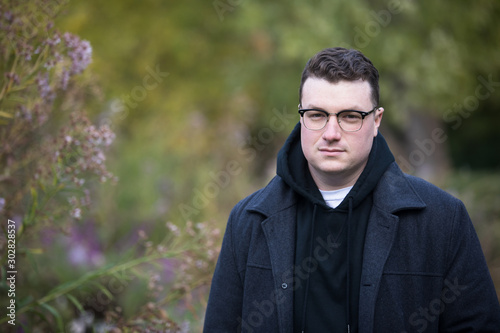 A caucasian male wearing a black hoodie sweater and glasses poses in a garden while surrounded by fall foliage on a cold afternoon.