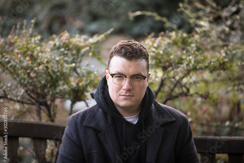A caucasian male wearing a black hoodie sweater and glasses poses in a garden while surrounded by fall foliage on a cold afternoon.