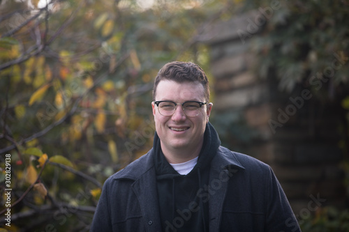 A caucasian male wearing a black hoodie sweater and glasses poses in a garden while surrounded by fall foliage on a cold afternoon.