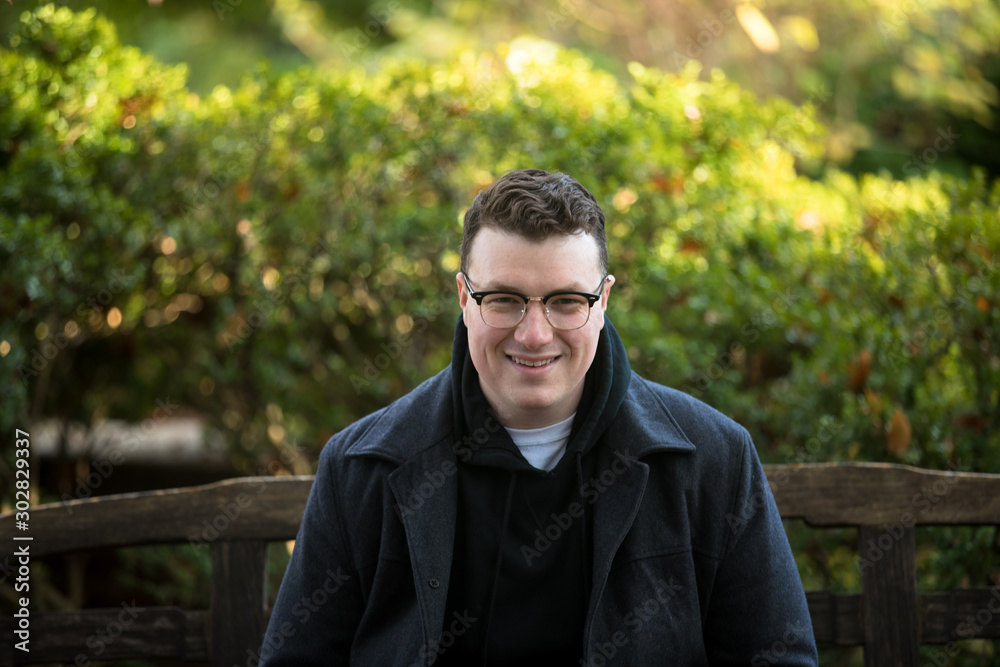 A caucasian male wearing a black hoodie sweater and glasses poses in a garden while surrounded by fall foliage on a cold afternoon.