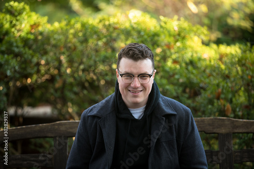 A caucasian male wearing a black hoodie sweater and glasses poses in a garden while surrounded by fall foliage on a cold afternoon.