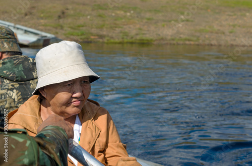 Sad adult Yakut woman in Panama looks at the river sitting in a boat with men on the brimstone. © Дмитрий Седаков