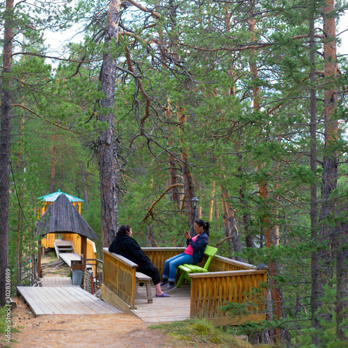Two Yakut girls are resting on a bench in the forest in the Park playing phone in the Northern taiga.