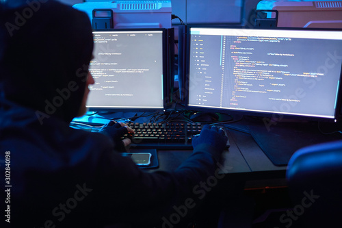 Rear view of computer hacker wearing hoody sitting at the table in front of computer monitors and breaking the computer codes in dark office