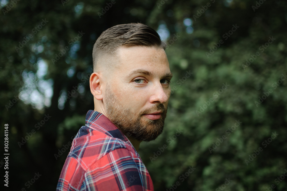 Brutal bearded young man in red shirt is standing outdoors on green trees background. Hipster style concept. Brutal bearded young man in red shirt is standing outdoors on green trees background. Hipster style concept.