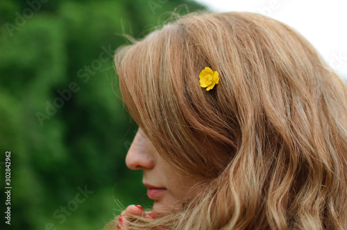 Outdoors portrait of young calm blonde woman in pink clothes with little yellow flower in her long hair on the hill with green forest background during spring or early autumn in the mountains