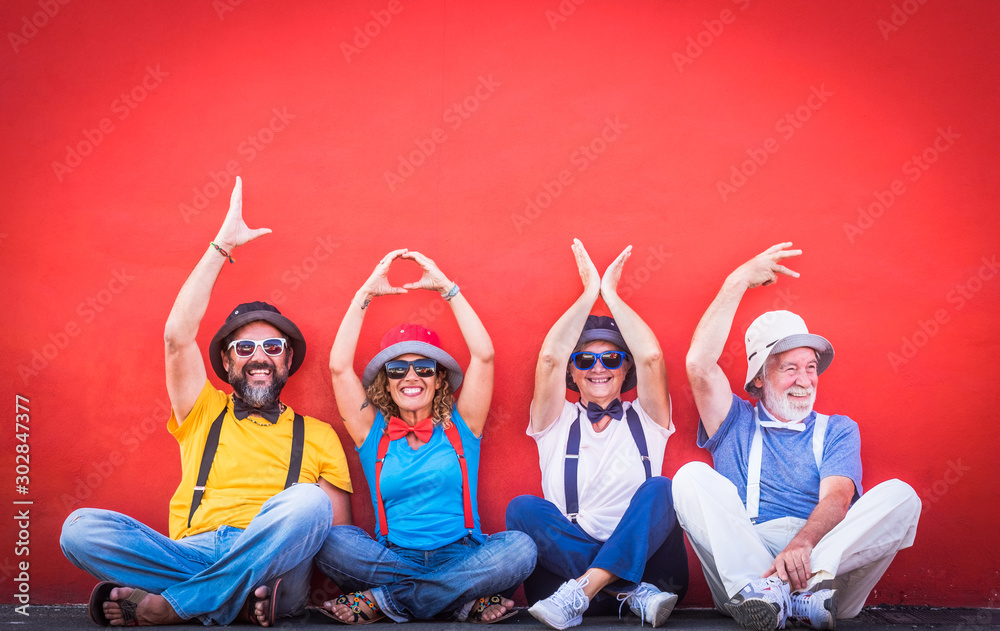 Fototapeta premium Happy family of four people sitting on the floor in front of a red wall. Gesturing with hands the word love. Large smiles and colorful dresses with suspenders and bow ties. Complicity and happiness