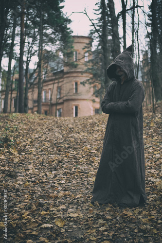 Obraz na plátně Mysterious monk, wizard or adept of secret society in hooded robe stands in forest against the background of medieval castle or monastery