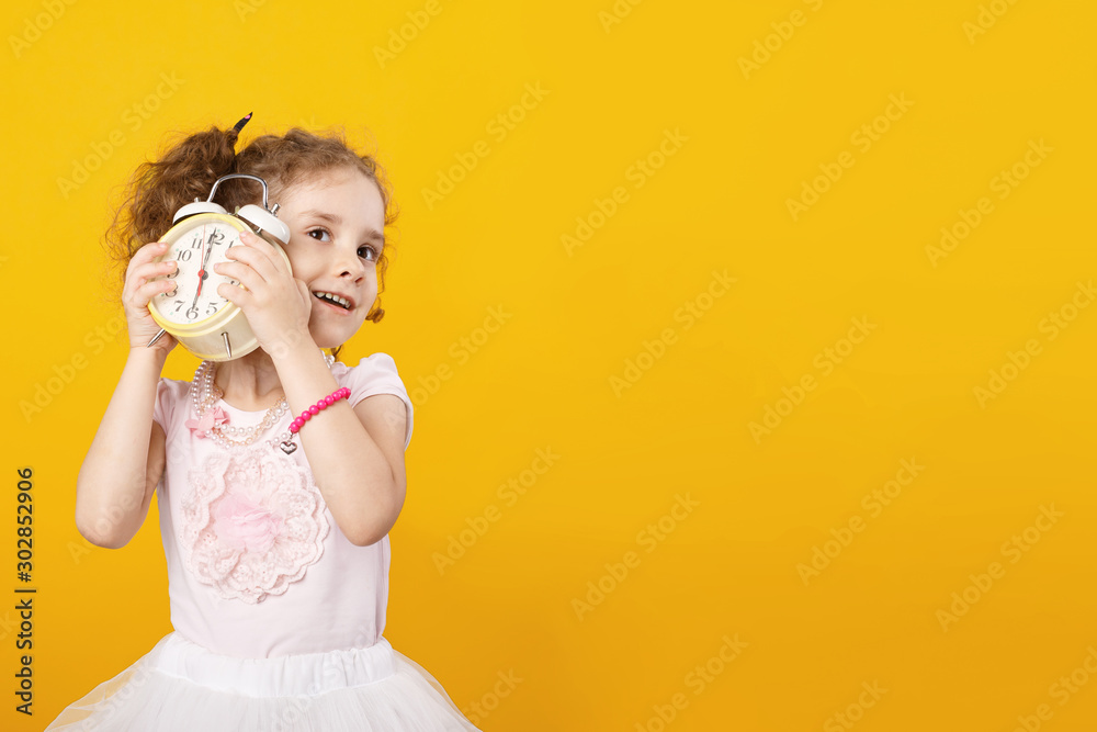 Little curiosity girl with curly hair holding near ears trying to listening a clock alarm, over yellow background.Space for text. Little curiosity girl with curly hair holding near ears trying to listening a clock alarm, over yellow background.Space for text.