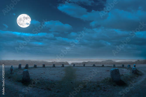 The mystical historic stone circle of Merry Maidens in Cornwall at night. It is foggy and the full moon is shining in the sky. Some clouds are in the sky.