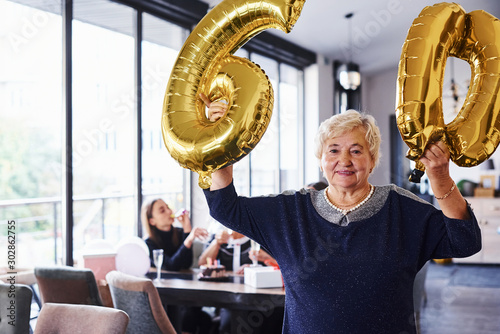 With balloons of number 60 in hands. Senior woman with family and friends celebrating a birthday indoors