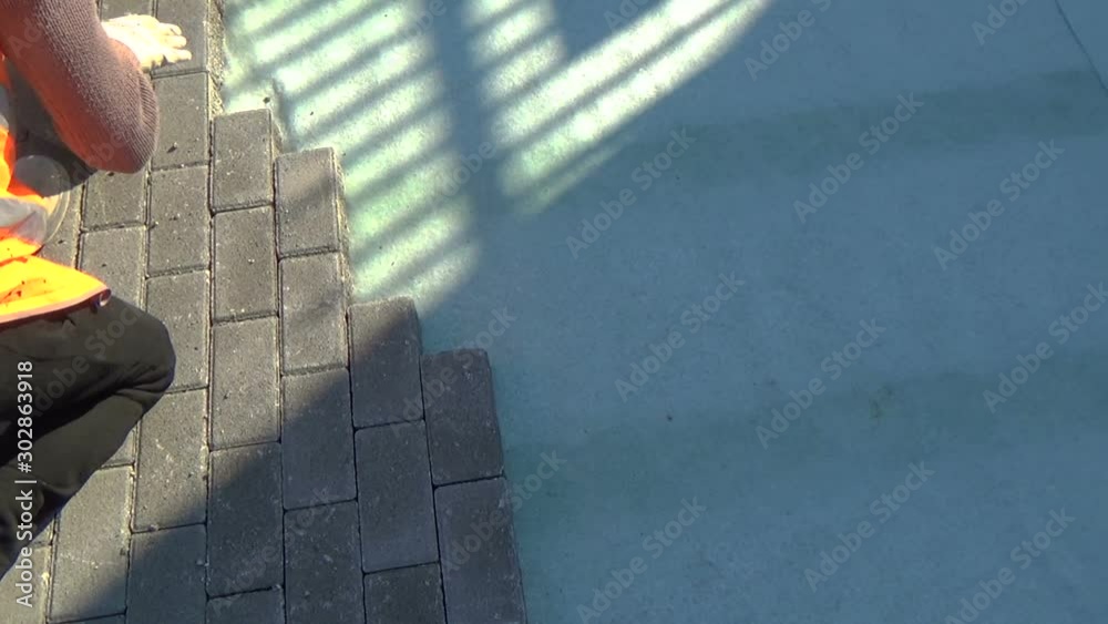 Construction worker laying interlocking paving concrete onto sheet nonwoven bedding sand and fitting them into place.