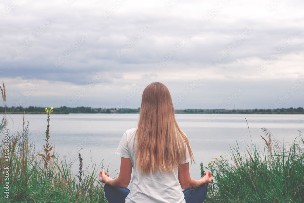 Beautiful girl with long hair is sitting on the shore. The view from the back. Sunset. Peace and tranquility. Yoga by the lake