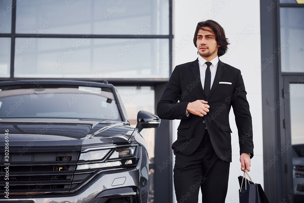 Portrait of handsome young businessman in black suit and tie outdoors near modern car and with shopping bags Portrait of handsome young businessman in black suit and tie outdoors near modern car and with shopping bags