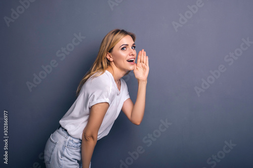 Caucasian woman in neutral casual outfit standing on a neutral grey background. Portrait with emotions: happiness, amazement, joy and satisfaction while whispering secrets