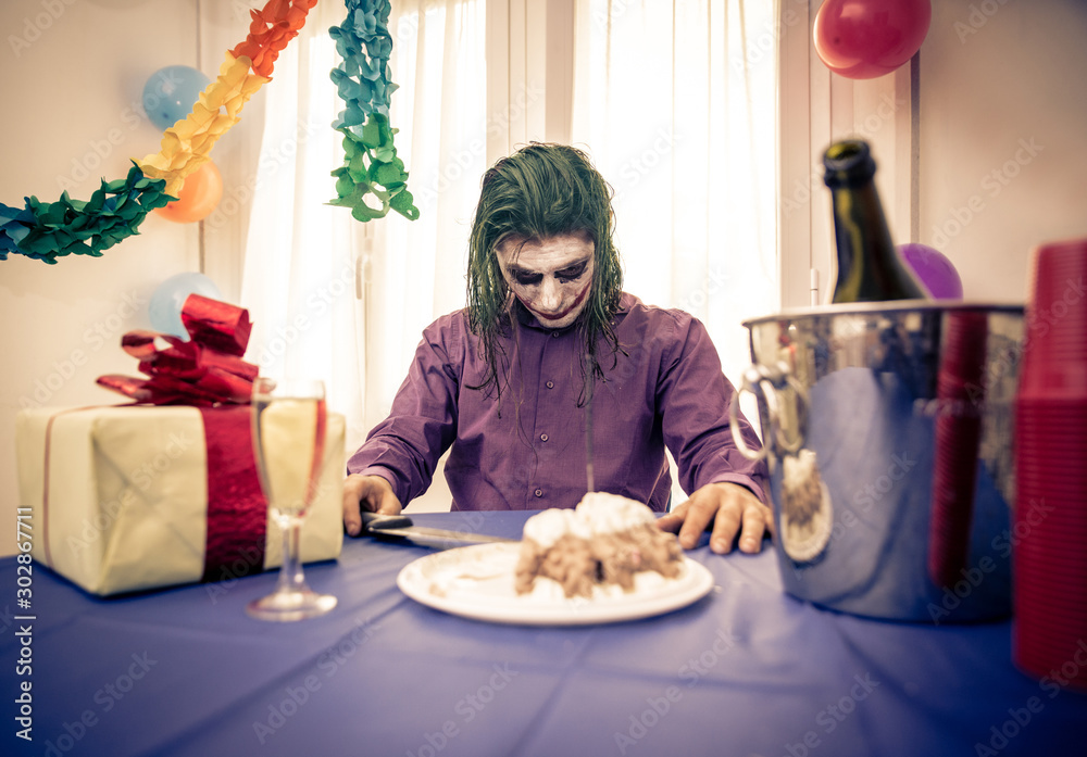 Clown celebrating birthday party alone at home. Sad man with mental disease sitting and eating cake фотография Stock