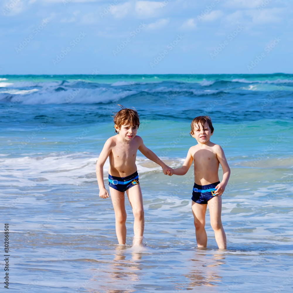 Two happy little kids boys running on the beach of ocean. Funny cute children, siblings, twins and best friends making vacations and enjoying summer day. Having fun with jumping into wave.