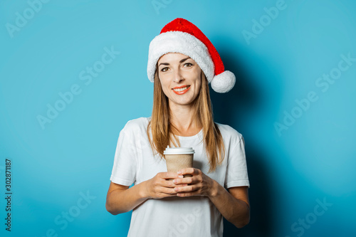 Beautiful Young woman with a smile in a hat of Santa Claus holds a cup of coffee on a blue background. Concept idea for New Year and Christmas  Christmas presents