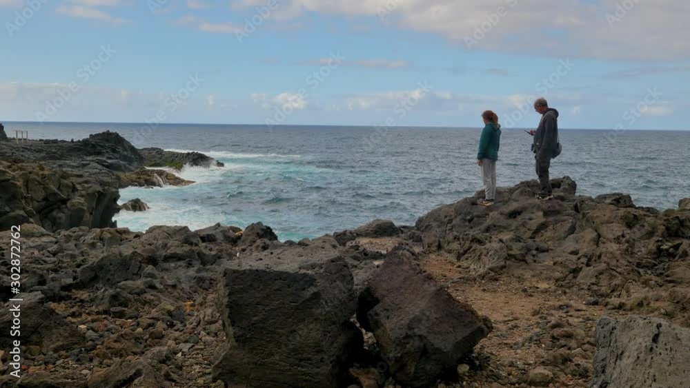 Handheld shot of an old man and daughter standing on a cliff over the waves hitting the rocky beach on an overcast day Handheld shot of an old man and daughter standing on a cliff over the waves hitting the rocky beach on an overcast day