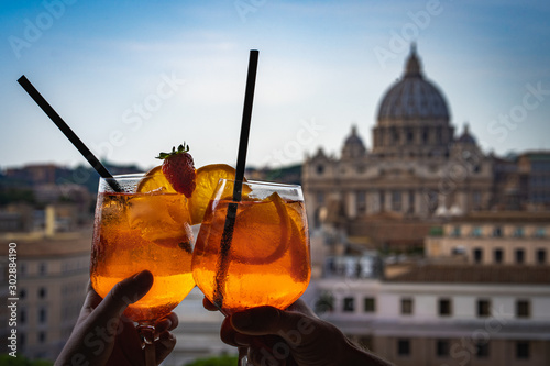 A couple is holding glasses of Aperol in the bar inside the Castle Sant'Angelo. Glasses of Aperol and St Peter's basilica at the background. Concept photo of traveling to Italy.