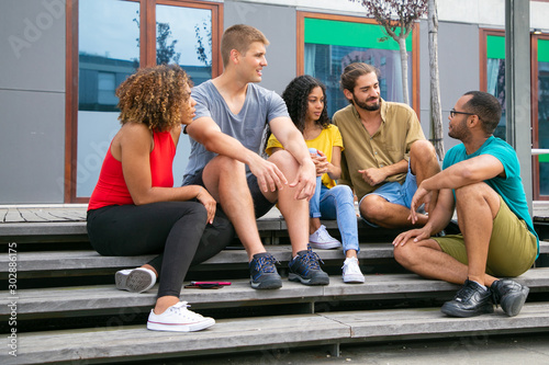 Young friends sitting on steps. Cheerful young multiethnic men and women sitting together on street and interacting. Friendship concept