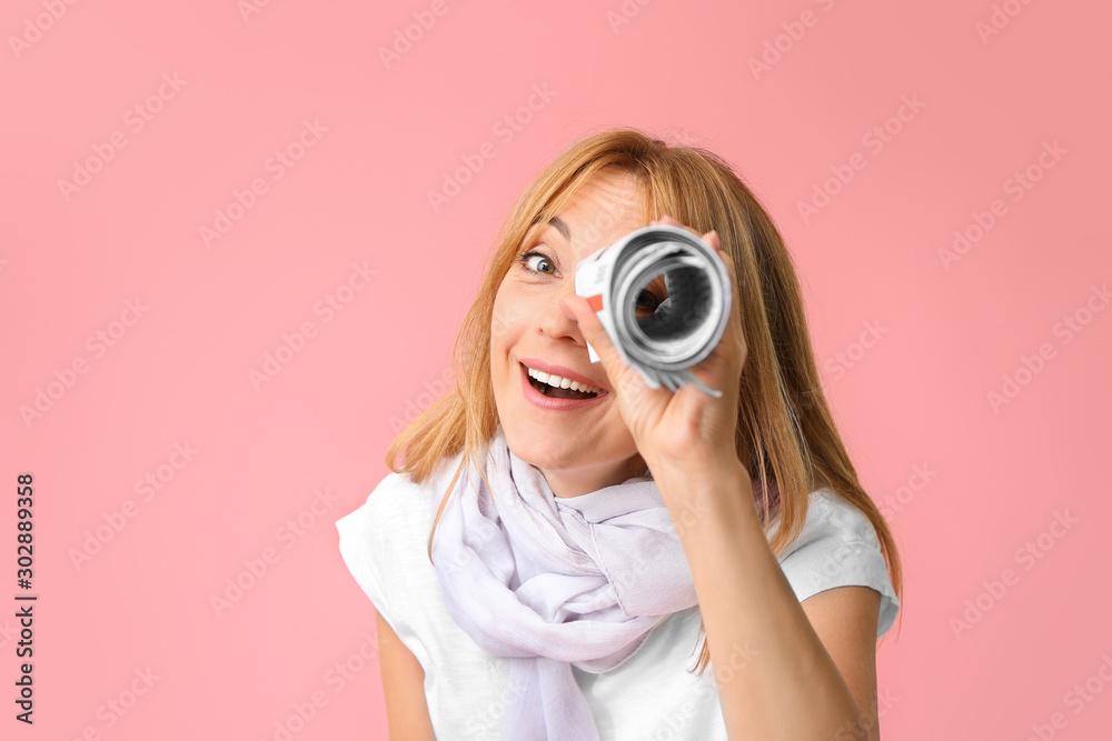 Fototapeta premium Stylish mature woman looking at viewer through rolled newspaper against color background