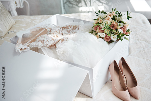 Luxury wedding dress in white box, beige women's shoes and bridal bouquet on bed, copy space. Bridal morning preparations. Wedding concept